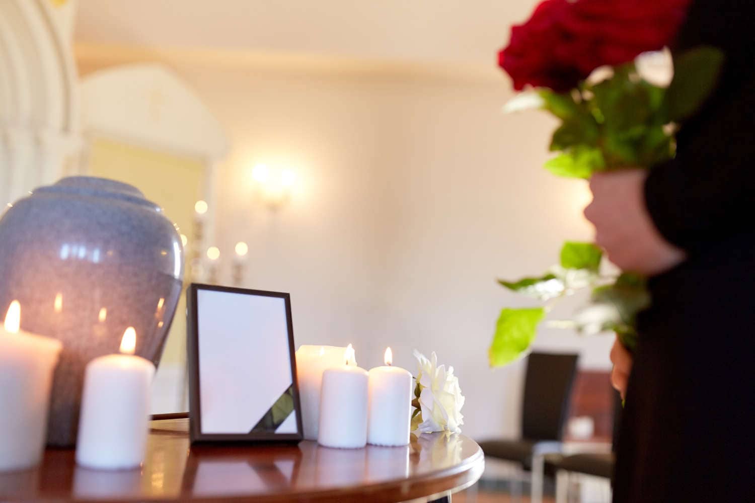 cremation, people and mourning concept -  cinerary urn, photo frame with black ribbon and woman holding red roses at funeral in church